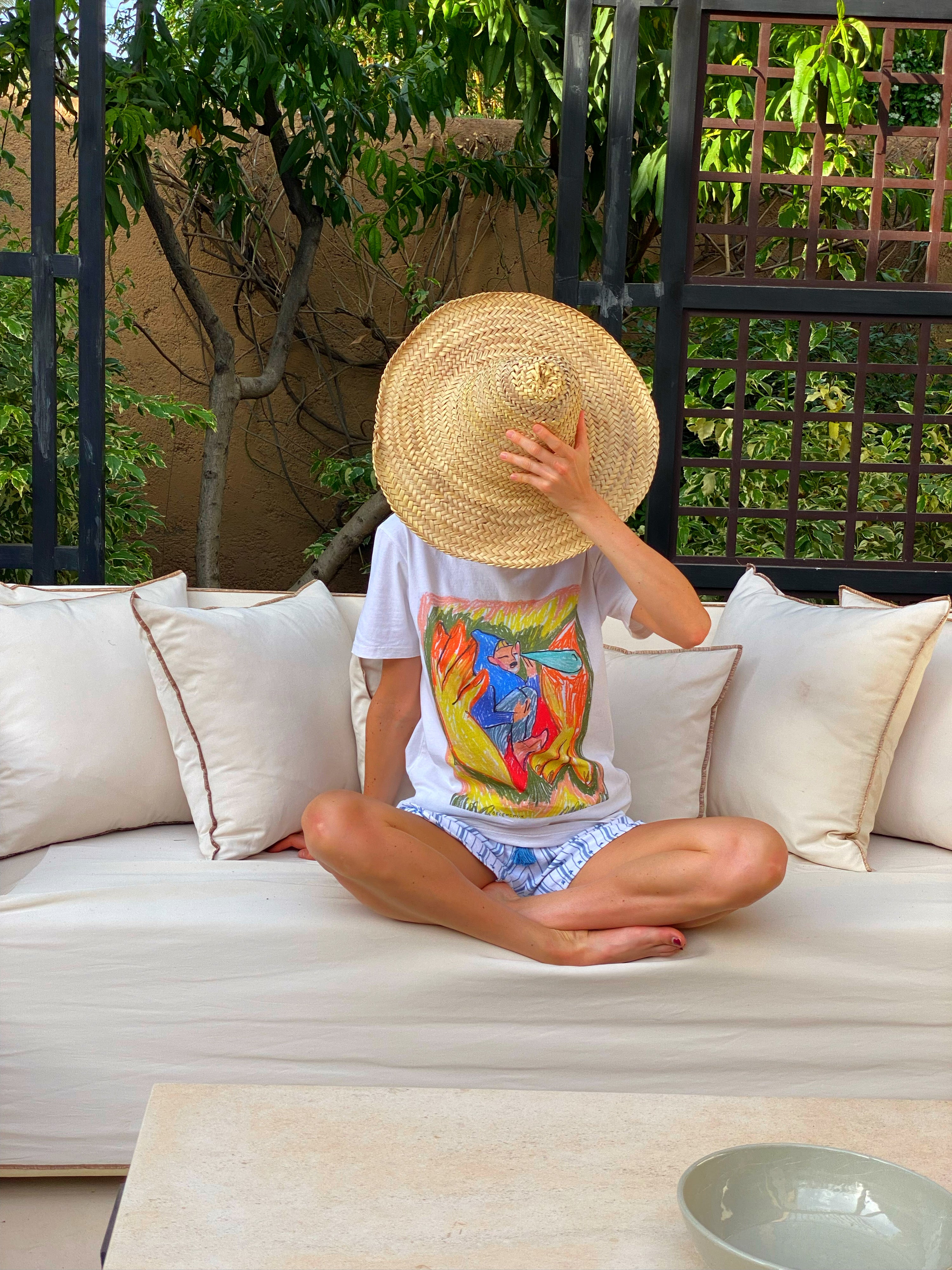 Person sitting on a couch with a colorful shirt and straw hat, surrounded by greenery.