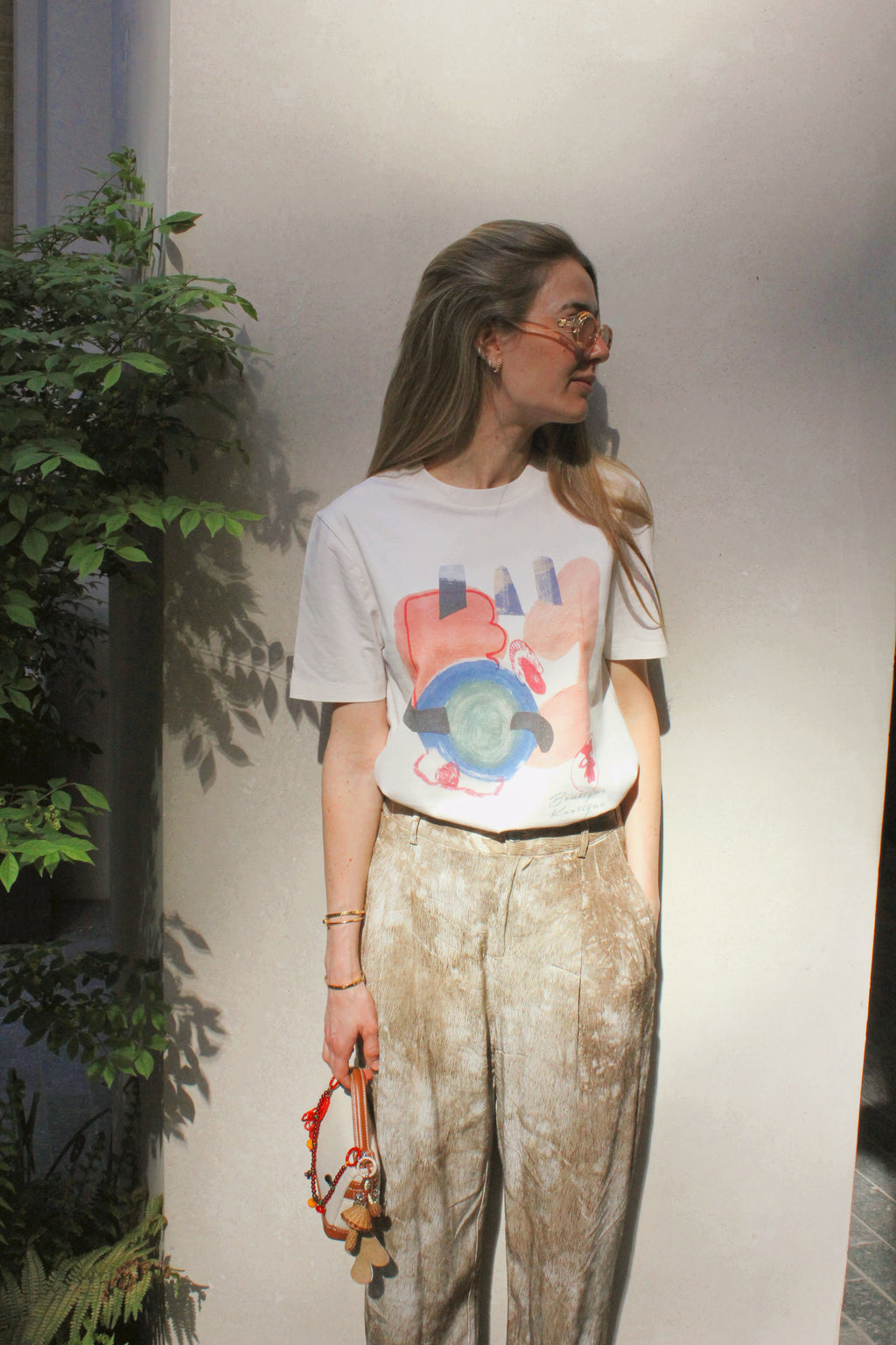 Person wearing a graphic t-shirt and light pants standing indoors with plants in the background. A white organic cotton t-shirt with a colorful abstract print and the text 'Breathe' on the front. My Brain is Offline tee — white organic cotton t-shirt with handwritten print, unisex fit, mental health charity