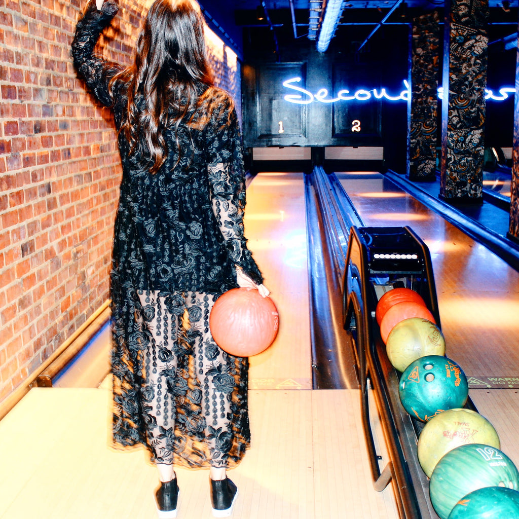 Woman holding a pink bowling ball at a bowling alley with colorful balls on the rack. Black lace dress — handmade black lace designer dress from Hanout Boutique Marrakech, fitted, limited edition occasion piece