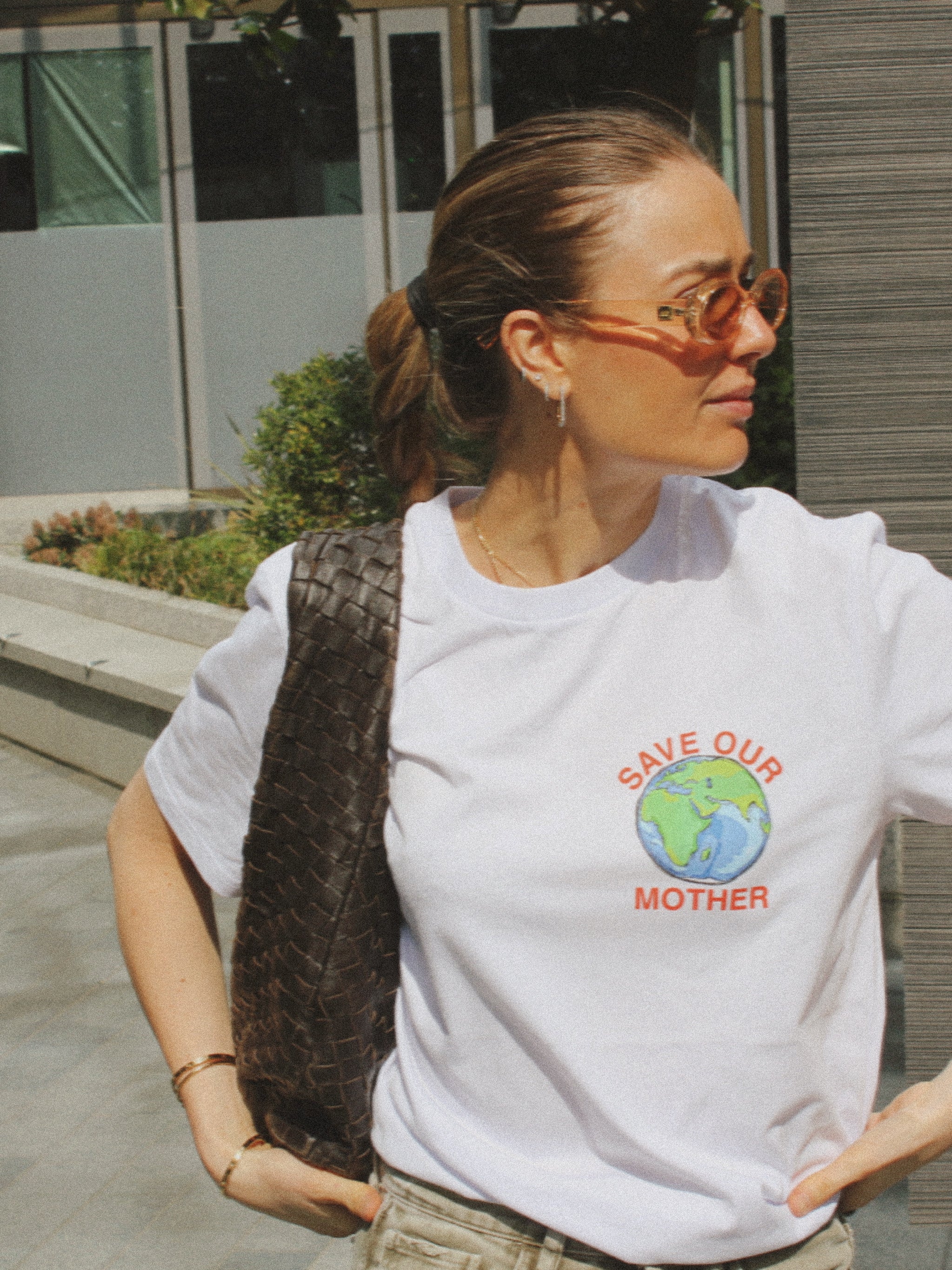 Save Our Mother climate tee — white organic cotton t-shirt with hand-illustrated earth print, unisex fit, climate charity tee. Girl in the city crossing the street with bottega bag