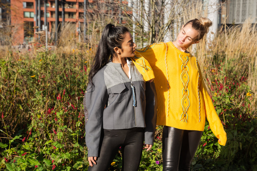 Two women standing outdoors in a garden setting, one wearing a gray jacket and black pants, the other in a yellow sweater with a pattern. Yellow bug wool jumper — handmade yellow wool designer jumper with bug print from Patrick Owen Jakarta, relaxed fit, limited edition