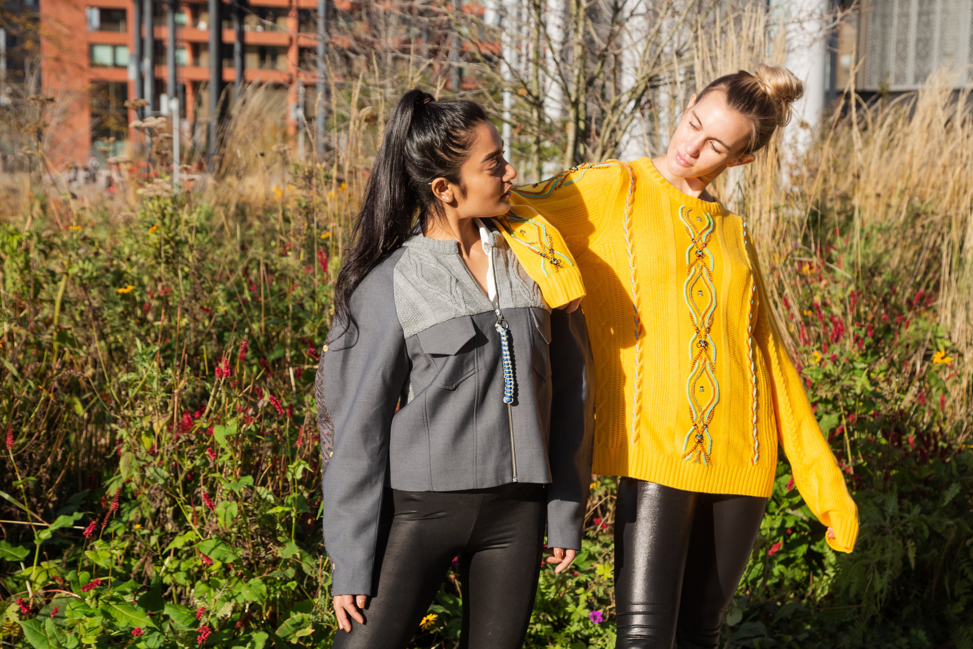 Two women standing outdoors in a garden setting, one wearing a gray jacket and black pants, the other in a yellow sweater with a pattern. Yellow bug wool jumper — handmade yellow wool designer jumper with bug print from Patrick Owen Jakarta, relaxed fit, limited edition