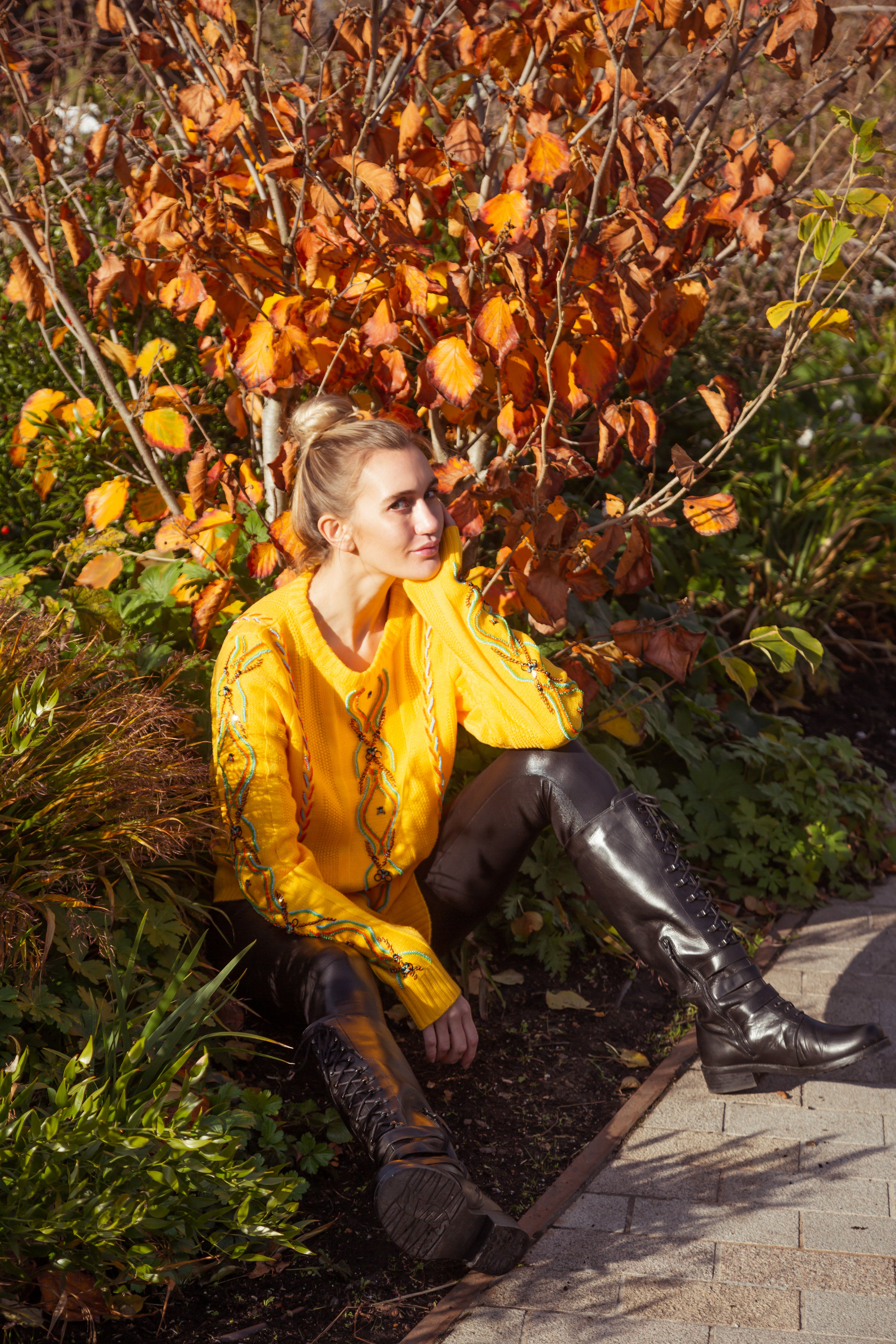 Woman in yellow coat and black boots sitting on a path with autumn foliage. Yellow bug wool jumper — handmade yellow wool designer jumper with bug print from Patrick Owen Jakarta, relaxed fit, limited edition