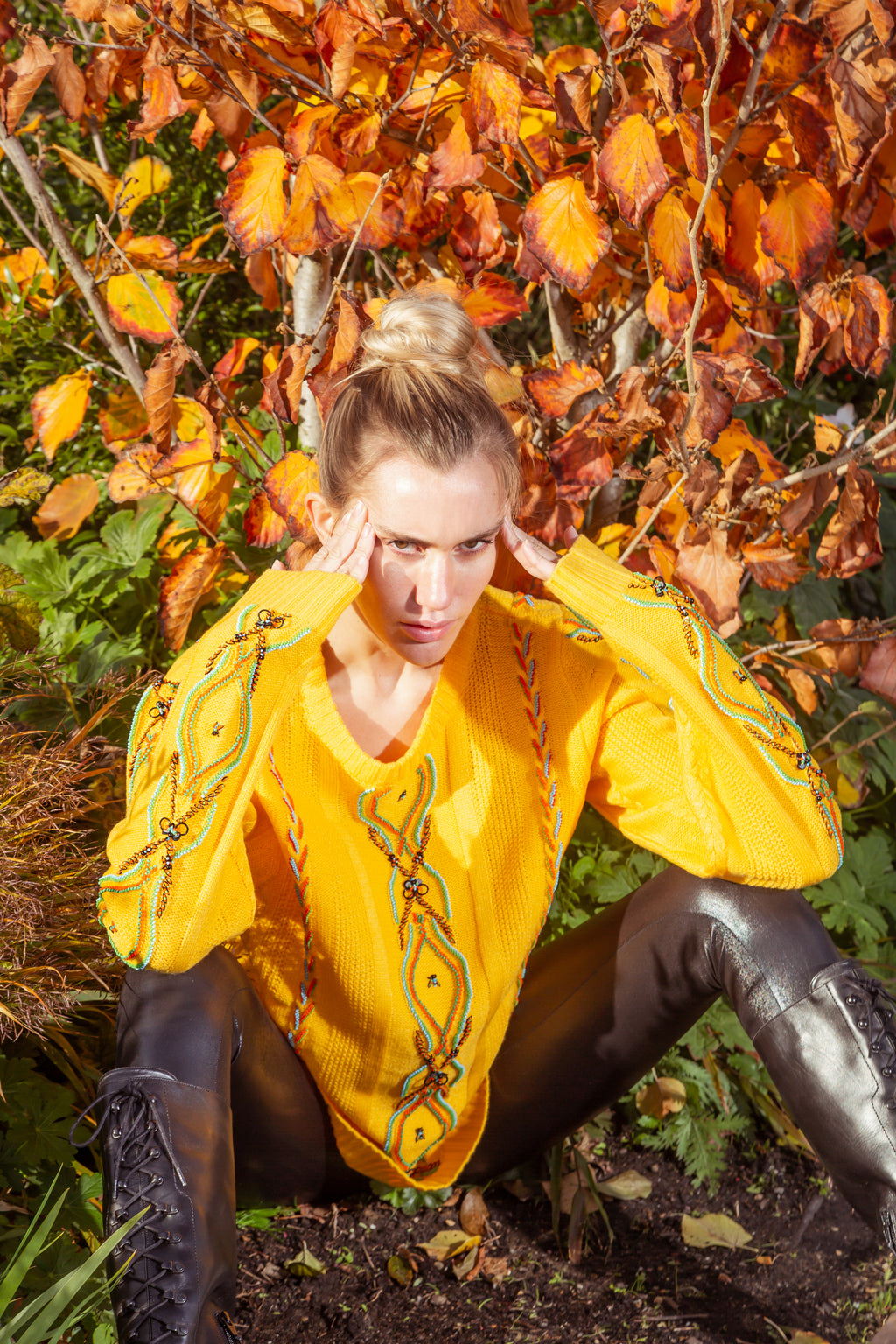 Woman in a yellow sweater and black pants sitting among autumn leaves. Yellow bug wool jumper — handmade yellow wool designer jumper with bug print from Patrick Owen Jakarta, relaxed fit, limited edition