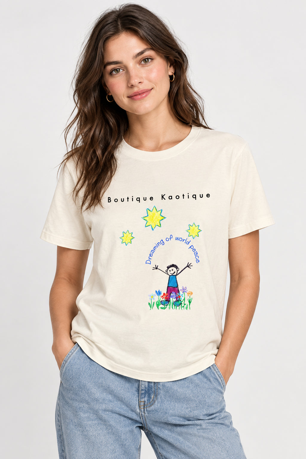 A cream-colored short-sleeve t-shirt with a graphic on the front. The graphic features the text 'Boutique Kaotique' and 'Dreaming of world peace' in blue, along with yellow starbursts, a cartoon child with outstretched arms, and small colorful flowers at the bottom.