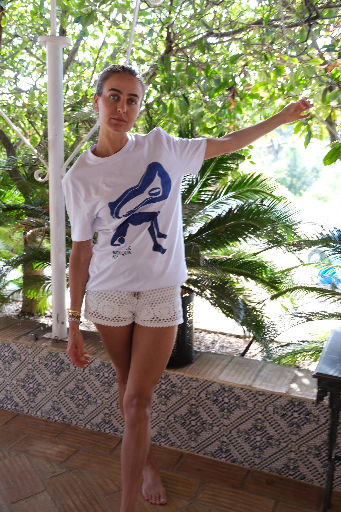 Person standing on a wooden floor with a white shirt and shorts, surrounded by greenery.