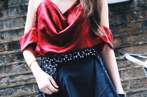 Person wearing a red top and dark skirt against a brick wall. Indonesian-inspired lace skirt — handmade lace designer skirt from Maryallé Jakarta, fitted, limited edition occasion piece. Cut out burgundy top — handmade burgundy designer top with cut-out detail from Maryallé Jakarta, fitted, limited edition