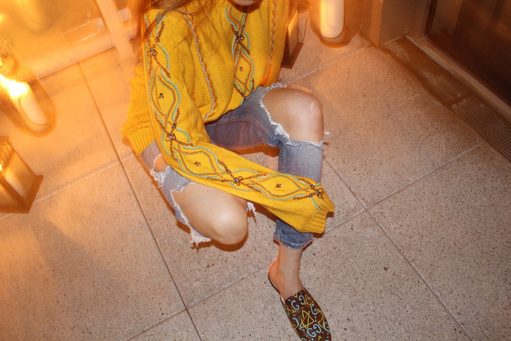 Person wearing a yellow patterned top and ripped denim shorts on a tiled floor. Yellow bug wool jumper — handmade yellow wool designer jumper with bug print from Patrick Owen Jakarta, relaxed fit, limited edition