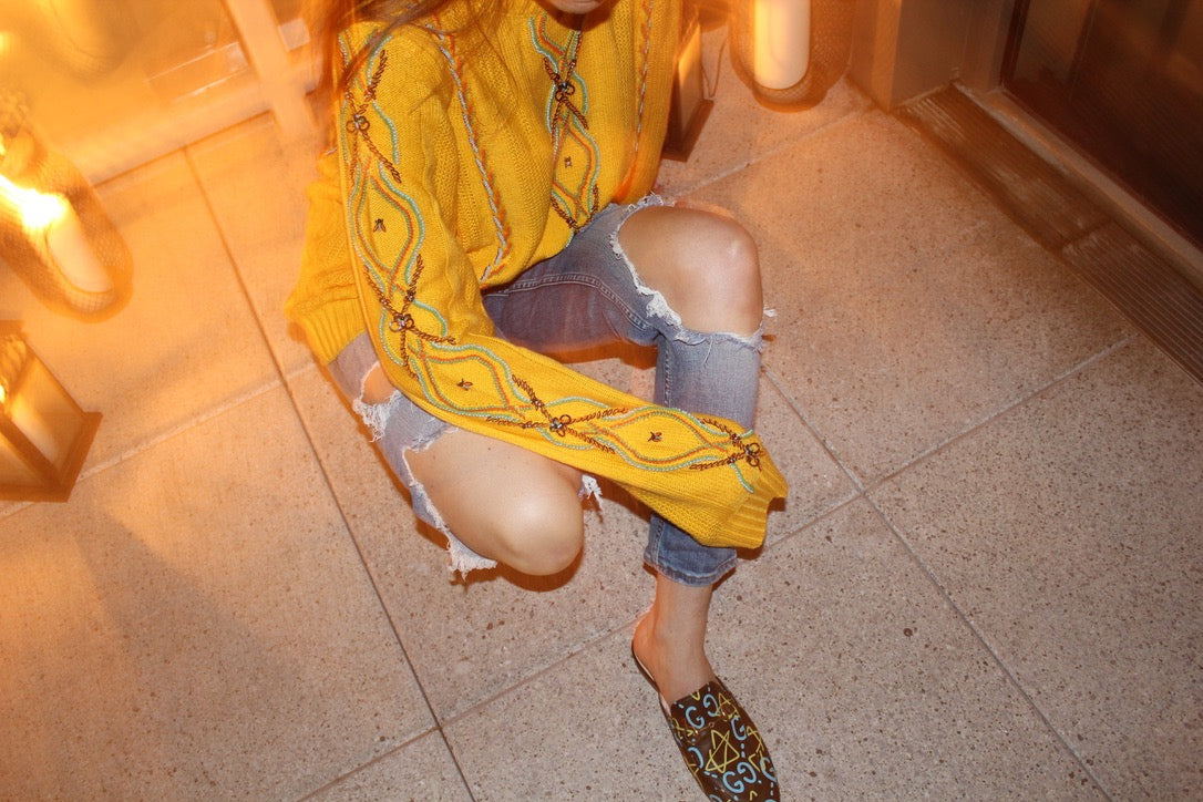 Person wearing a yellow patterned top and ripped denim shorts on a tiled floor. Yellow bug wool jumper — handmade yellow wool designer jumper with bug print from Patrick Owen Jakarta, relaxed fit, limited edition