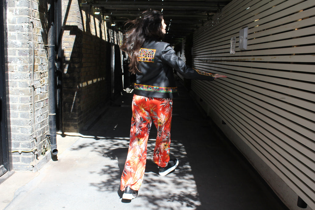 Person wearing a black jacket with colorful pants walking through an alleyway. Orange patterned silk pants — handmade orange silk designer pants with vibrant print from Hanout Boutique Marrakech, relaxed fit