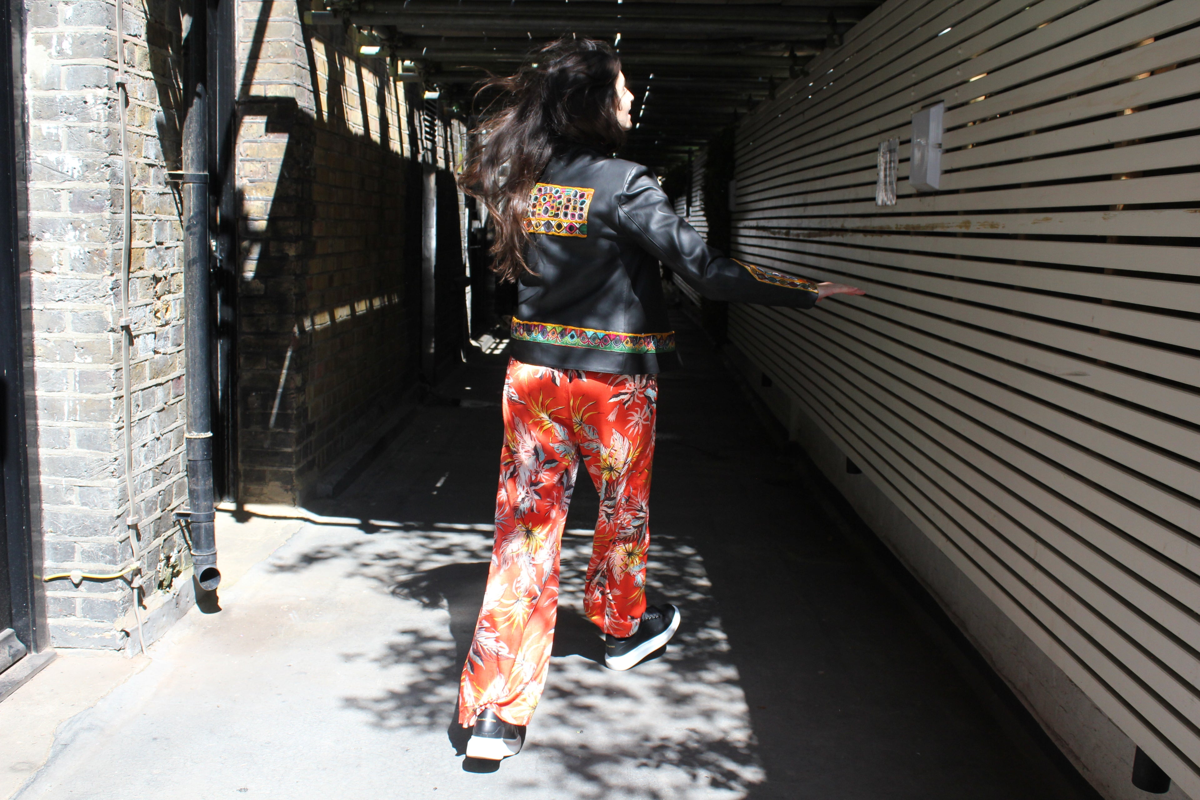 Person wearing a black jacket with colorful pants walking through an alleyway. Orange patterned silk pants — handmade orange silk designer pants with vibrant print from Hanout Boutique Marrakech, relaxed fit
