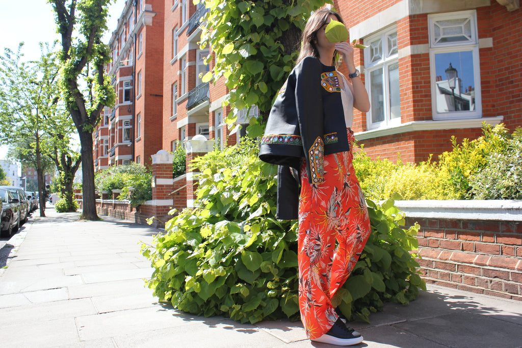 Woman in a colorful clothing standing on a street with brick buildings and greenery. Orange patterned silk pants — handmade orange silk designer pants with vibrant print from Hanout Boutique Marrakech, relaxed fit