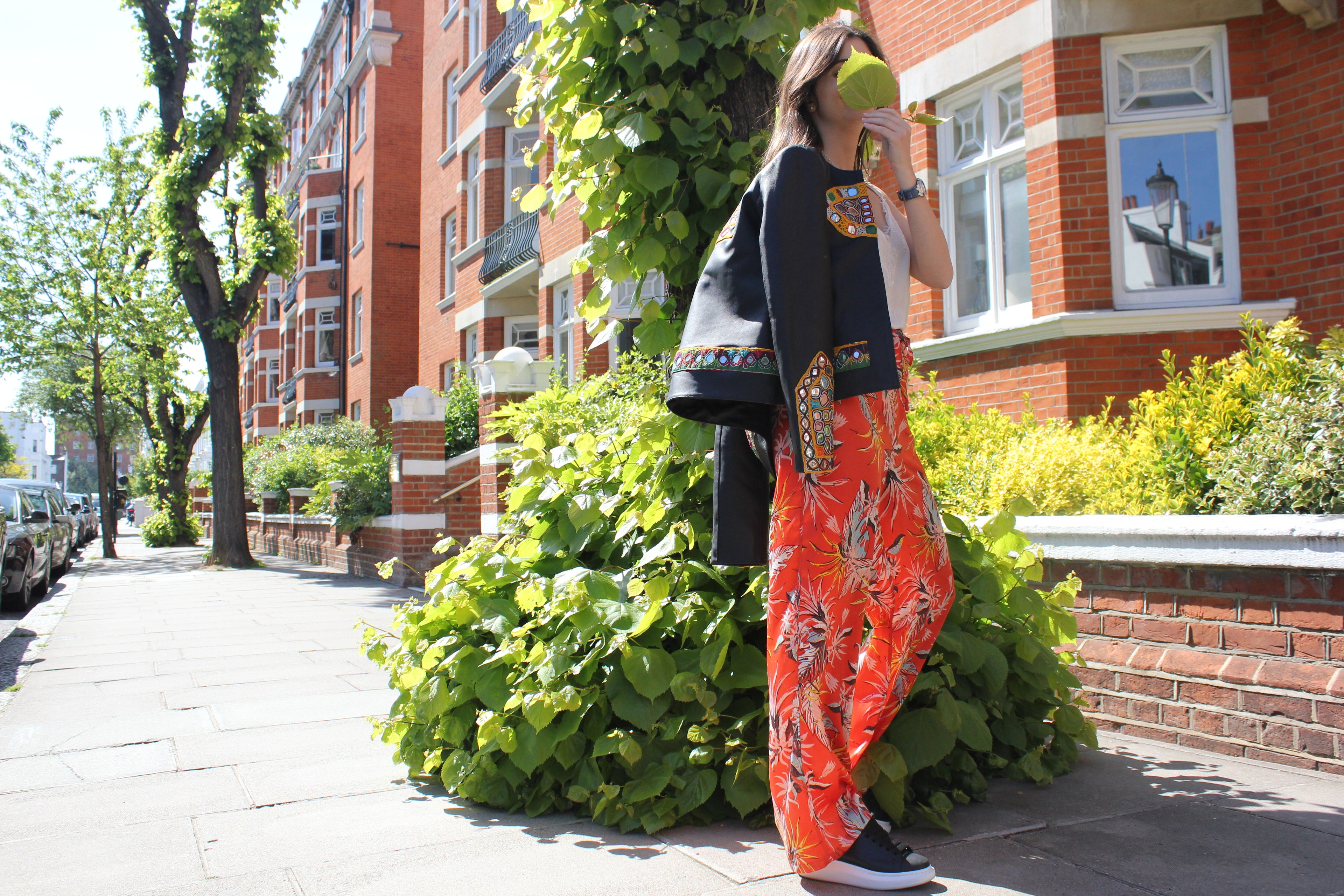 Woman in a colorful clothing standing on a street with brick buildings and greenery. Orange patterned silk pants — handmade orange silk designer pants with vibrant print from Hanout Boutique Marrakech, relaxed fit