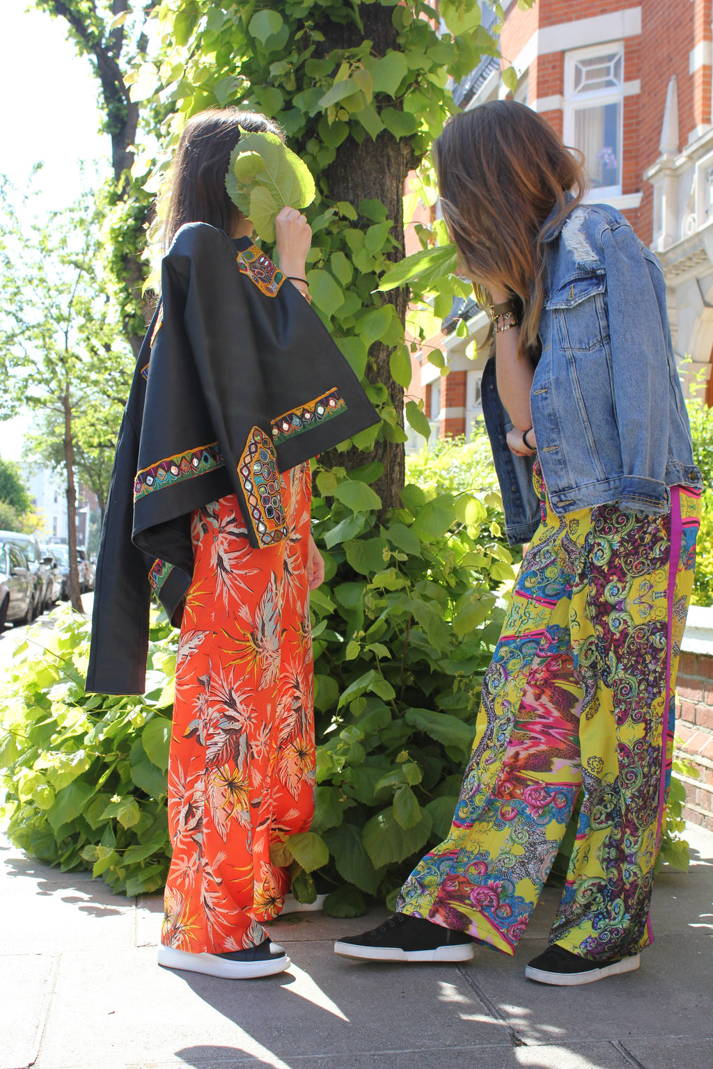 Person wearing colorful floral-patterned pants and a black jacket in an outdoor setting. Orange patterned silk pants — handmade orange silk designer pants with vibrant print from Hanout Boutique Marrakech, relaxed fit