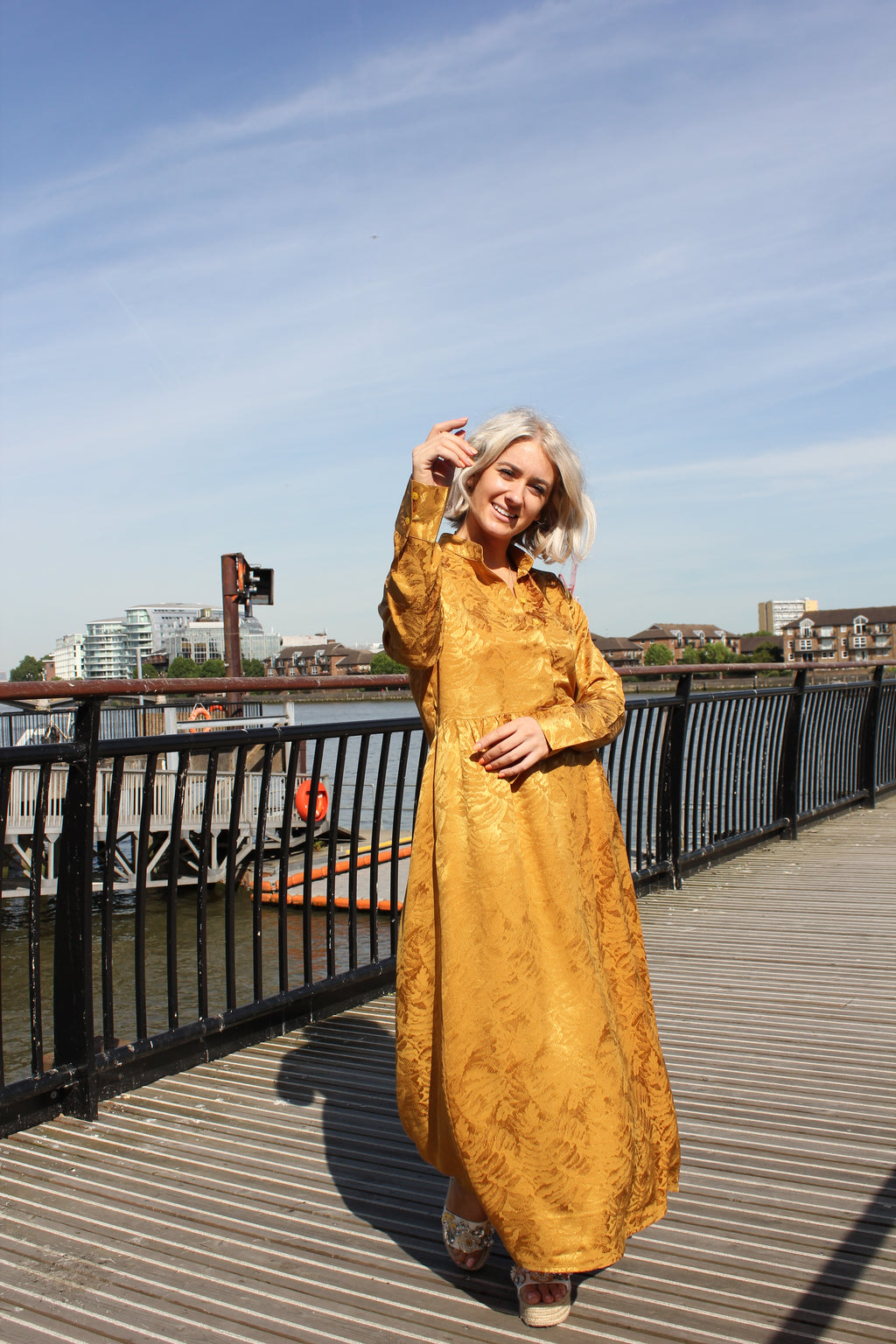 Person wearing a long, patterned dress standing on a rooftop with a cityscape in the background. Golden Hour dress — handmade yellow gold designer dress from Hanout Boutique Marrakech, fitted, limited edition occasion piece