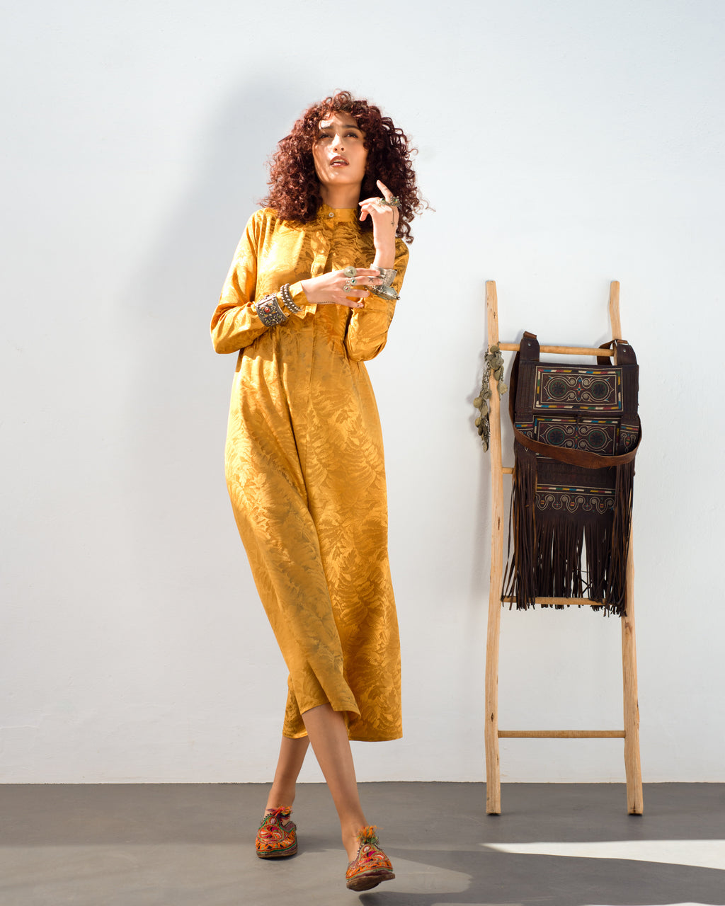 Moroccan Woman in a mustard yellow gold dress standing next to a wooden ladder with bags on it against a white wall. Golden Hour dress — handmade yellow gold designer dress from Hanout Boutique Marrakech, fitted, limited edition occasion piece