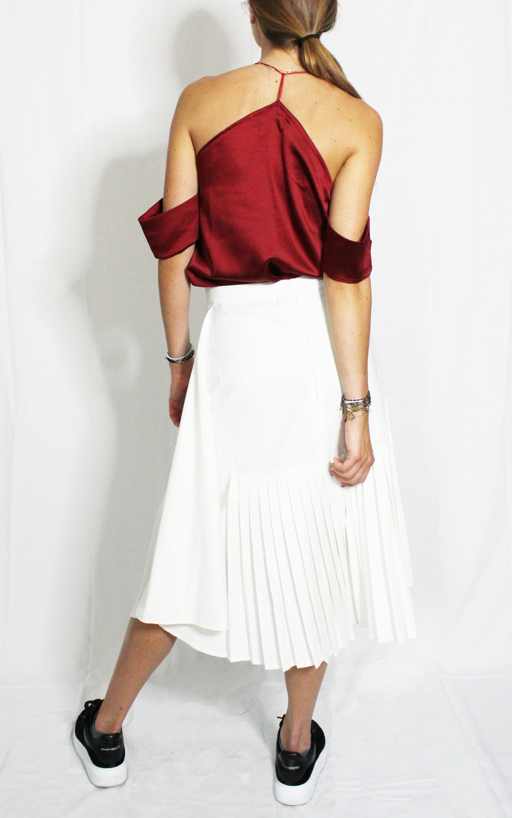 Person wearing a burgundy top and white pleated skirt on a plain background. Indonesian asymmetrical skirt — handmade asymmetric designer skirt from Major Minor Jakarta, relaxed fit, limited edition