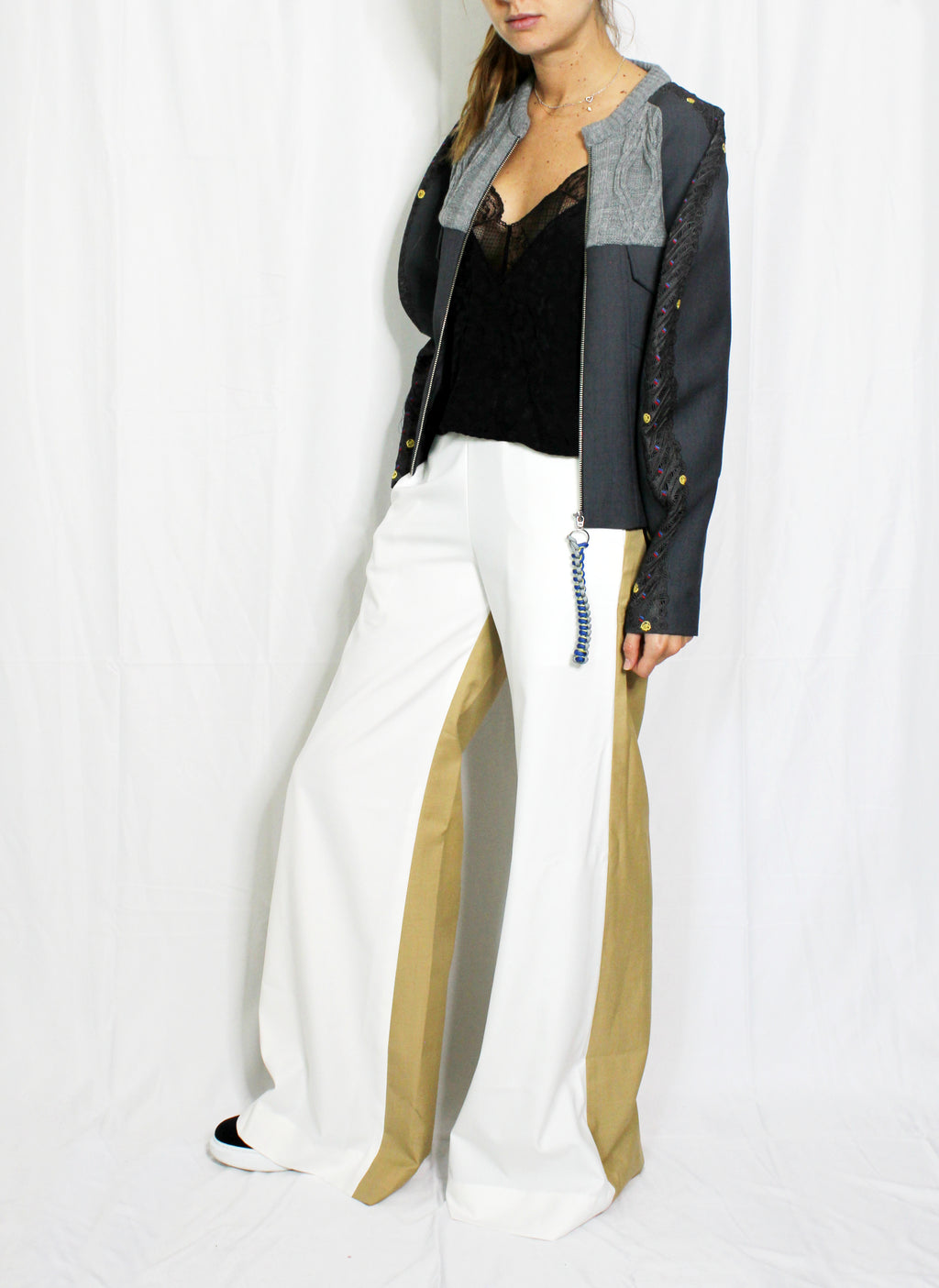Person wearing a black jacket with geometric patterns, black top, white pants with gold accents, and white shoes on a white background. Knitted bomber jacket — handmade designer knitted bomber from Patrick Owen Jakarta, relaxed fit, limited edition winter piece. Double coloured trousers — handmade designer two-tone trousers from Major Minor Jakarta, relaxed fit, limited edition piece
