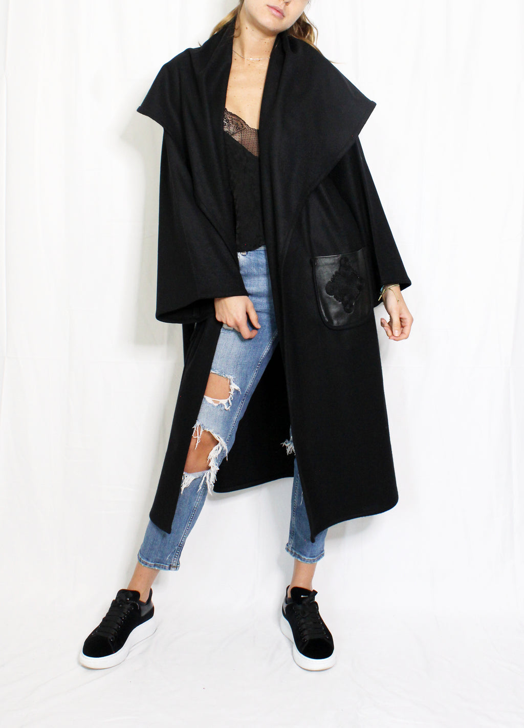 Person wearing a black coat, black top, and ripped jeans on a white background. Wool embroidered coat — heavy wool designer coat with hand-embroidered detail from Hanout Boutique Marrakech, limited edition