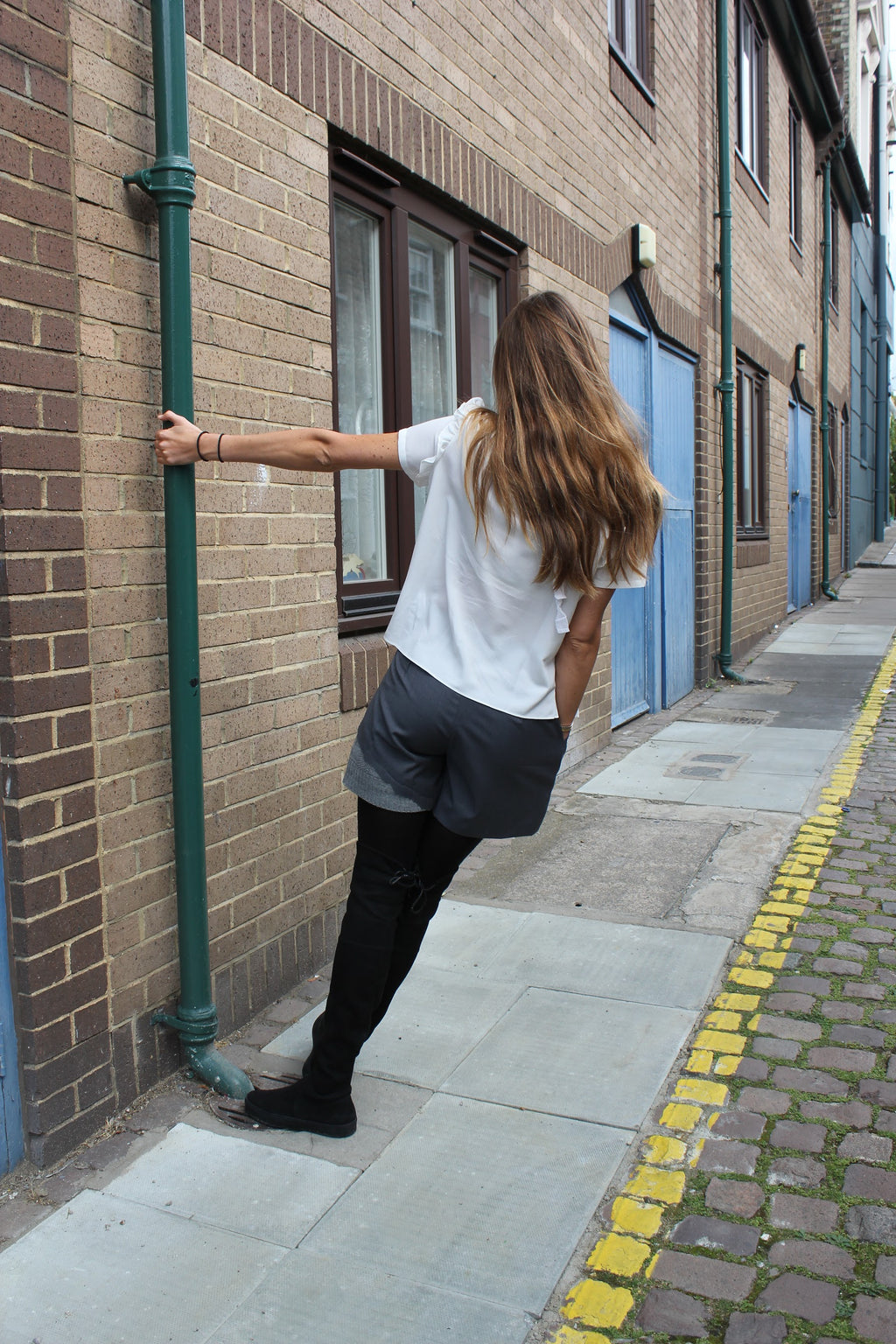 Person leaning against a brick wall on a street with cobblestone pavement. Wool blend shorts — handmade designer wool blend shorts from Patrick Owen Jakarta, tailored fit, limited edition piece