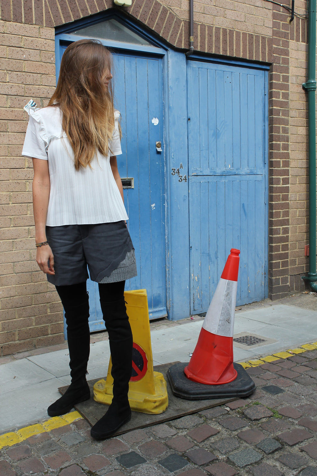 Person standing on a street with a blue door and traffic cones in the background. Wool blend shorts — handmade designer wool blend shorts from Patrick Owen Jakarta, tailored fit, limited edition piece