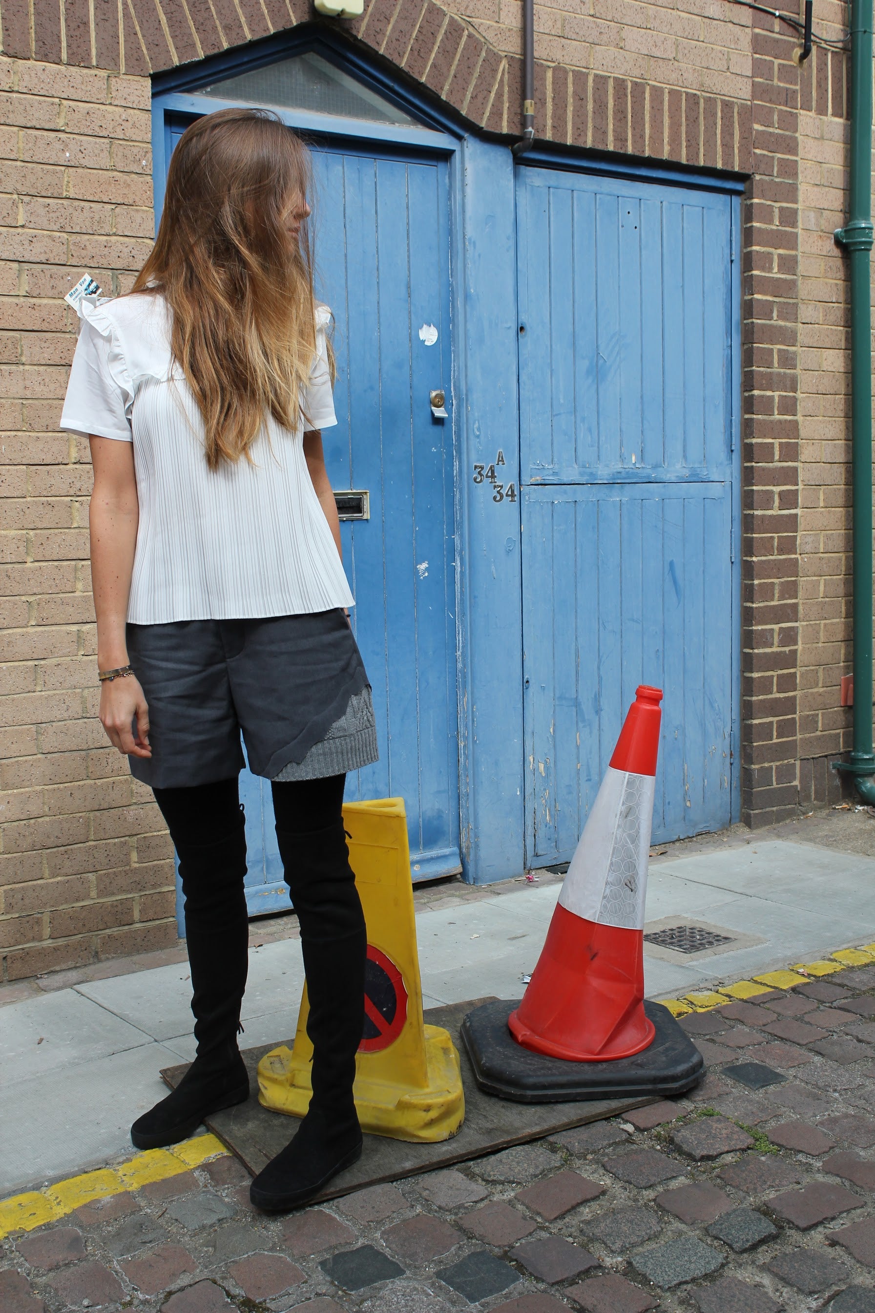 Person standing on a street with a blue door and traffic cones in the background. Wool blend shorts — handmade designer wool blend shorts from Patrick Owen Jakarta, tailored fit, limited edition piece