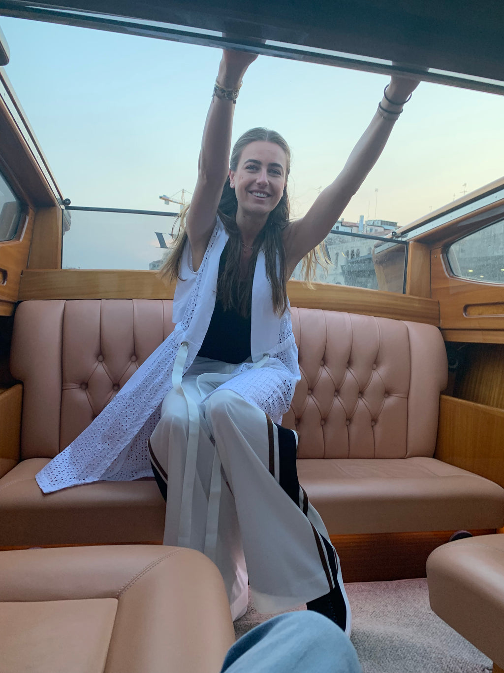 Woman sitting in a boat with beige interior, raising her arms in the air. Indonesian cool summer vest — handmade designer cotton summer vest from Major Minor Jakarta, relaxed fit, limited edition