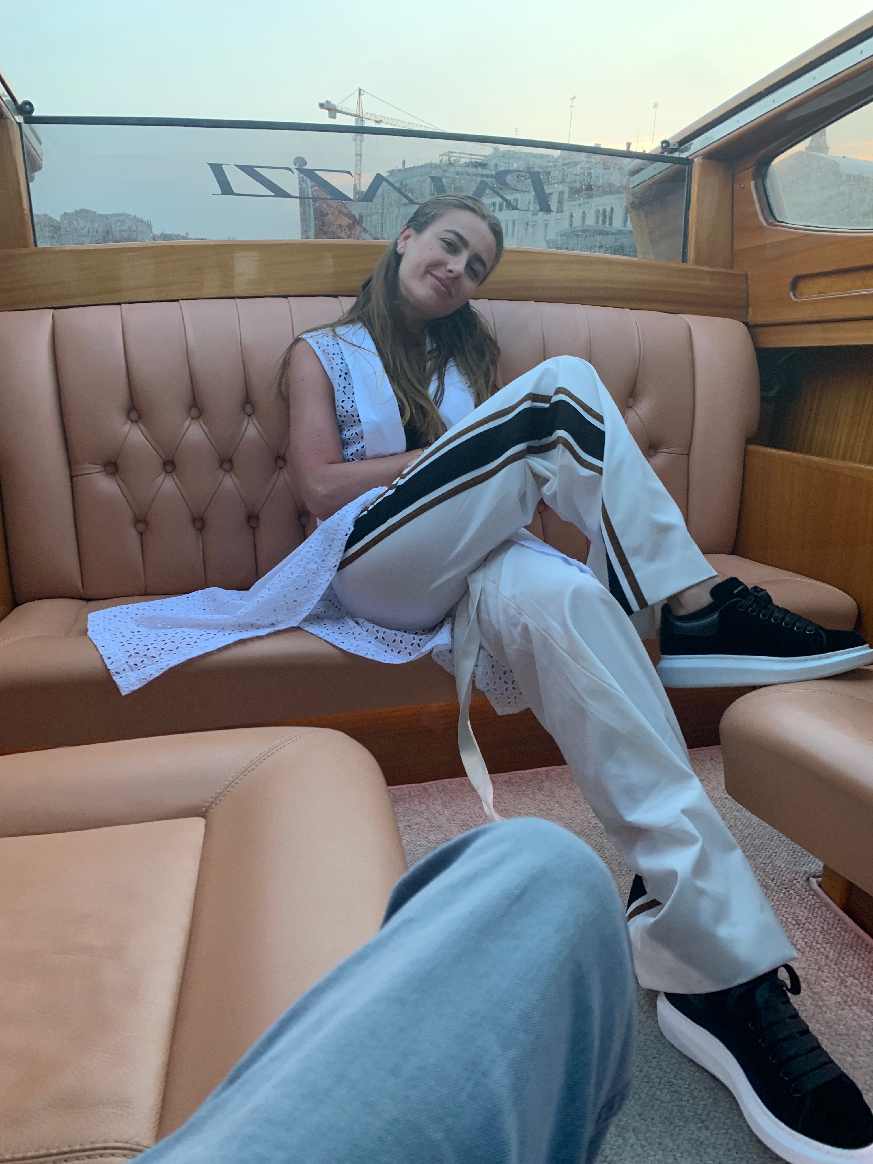 Person sitting on a boat with beige interior, wearing white pants and black shoes. Indonesian cool summer vest — handmade designer cotton summer vest from Major Minor Jakarta, relaxed fit, limited edition