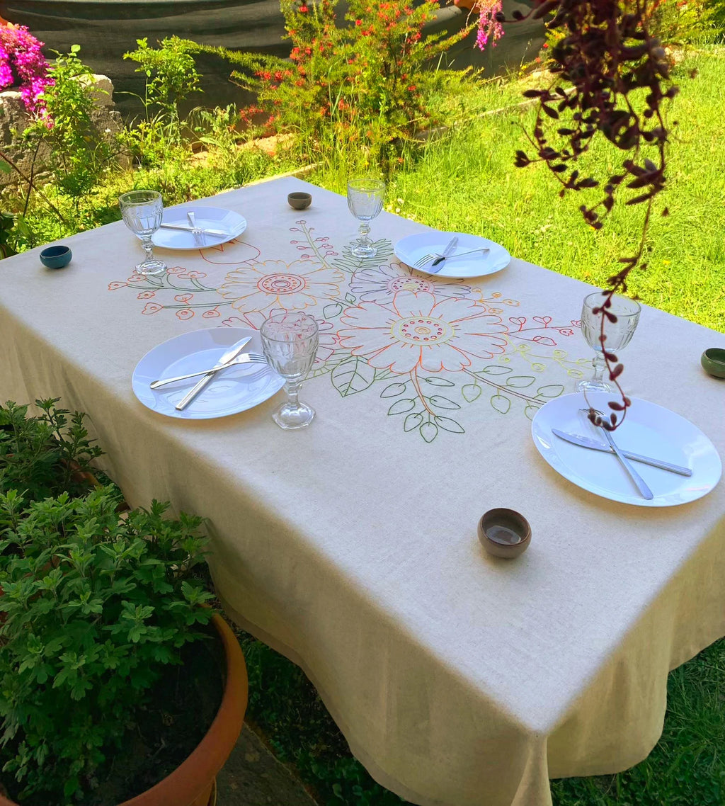 Dining table set with plates, glasses, and cutlery on a decorative tablecloth outdoors. Colorful floral embroidery design on a light background tablecloth